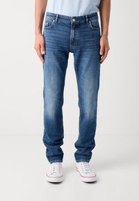 Klassiske blå denimjeans med rett benpassform, middels vask, subtil falming, fem lommer, glidelås og knappelukking, brukt med hvite sneakers.