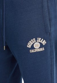 Tamsiai mėlynos šortai pagaminti iš minkštos medžiagos, su tekstūruotu ženkleliu, ant kurio yra "GUESS JEANS CALIFORNIA" logotipas kremine ir auksine siuvinėjimu.