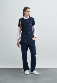 Laivastonsininen palmikkoneule vaaleanpunaisen kauluspaidan päällä, yhdistetty tummiin farkkuihin ja valkoisiin lenkkareihin. Ruskea olkalaukku.