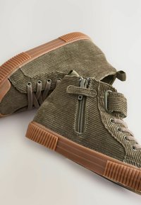Baskets montantes en velours côtelé vert olive avec une finition texturée, semelles en caoutchouc, fermeture éclair sur le côté et design à lacets avec œillets en métal.