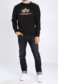 Mies, joka käyttää mustaa Alpha Industries -collegepaitaa, tummia käärittyjä farkkuja ja valkoisia lenkkitossuja, seisoo yhdellä kädellä taskussa.