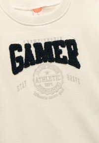 Vit sweatshirt med texturerad mörk marinblå "GAMER"-text, en rund, ljusgrå sportlogga och texten "STAY BRAVE". Mjuk tyg.