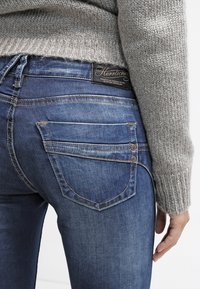 Person som bär blå denimjeans och en grå stickad tröja, som visar bakfickan och en klädlapp ovanför midjan.