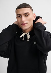 Svart hoodie med dragsko, med en liten logotyp på bröstet. Tyget verkar slätt och mjukt, med en avslappnad passform.