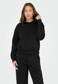 Giovane donna con capelli lunghi, che indossa un cappuccio nero e pantaloni neri, mentre tiene il laccio del cappuccio con una mano.