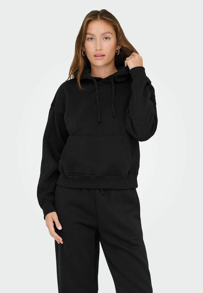 Giovane donna con capelli lunghi, che indossa un cappuccio nero e pantaloni neri, mentre tiene il laccio del cappuccio con una mano.