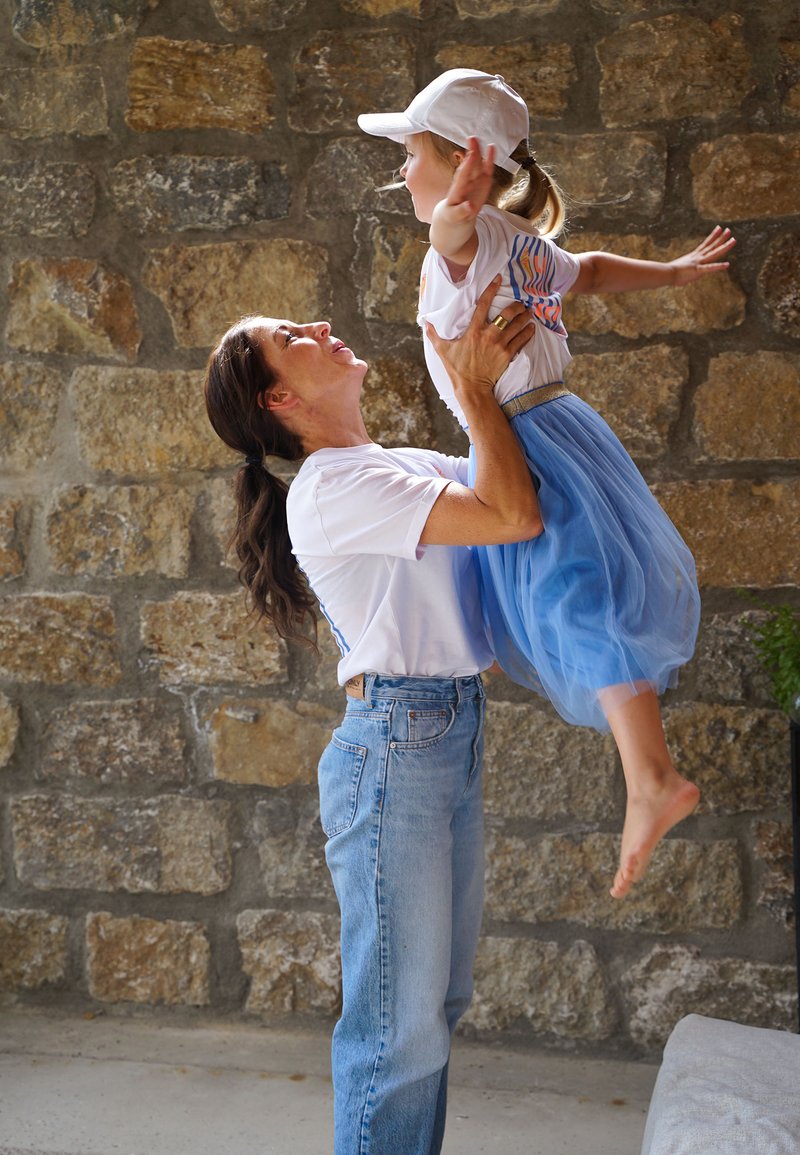 Frau in weißem Hemd und Jeans hebt ein junges Mädchen in blauem Rock und weißer Kappe hoch, beide lächeln vor einem Hintergrund aus Steinmauer.