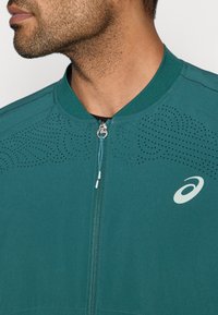 Αθλητικό τζάκετ σε τιρκουάζ χρώμα με φερμουάρ στο μπροστινό μέρος, ριμπ γιακά και διάτρητες λεπτομέρειες στους ώμους. Διαθέτει λογότυπο στο αριστερό στήθος.