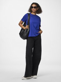 Kvinna klädd i svarta jeans med vida ben, blå T-shirt, svarta solglasögon, svart axelväska och svartvita sneakers som står mot en vit bakgrund.