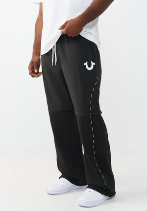 Man draagt gelaagde zwarte shorts over een zwarte broek met wit logo op de shorts en witte sneakers, staand tegen een effen witte achtergrond.