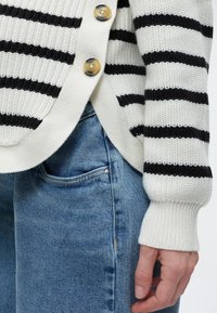 Približeni prikaz ruke blizu džepa plavih traperica, u bijelom pletenom kardiganu s crnim horizontalnim prugama i dva velika gumba.