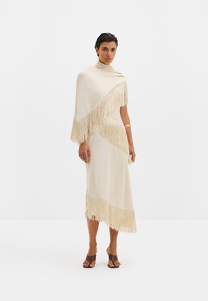 Frau mit kurzen dunklen Haaren, die einen cremefarbenen Fransen-Schal und ein passendes midilanges Kleid trägt, braune offene Schuhe mit Absatz und ein goldenes Armband.