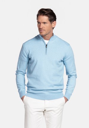 Man draagt een lichtblauwe trui met een korte ritssluiting over een wit overhemd en een witte broek, staand met handen in de zakken tegen een effen achtergrond.