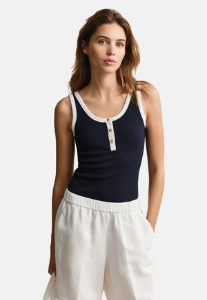 Vrouw die een nachtblauwe mouwloze top met witte bies en knopen draagt, gecombineerd met losse witte broek, staand tegen een effen achtergrond.