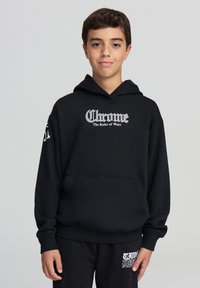 Ragazzo che indossa un hoodie nero con la scritta "Chrome The Ruler of Ways" e pantaloni della tuta neri abbinati, in piedi contro uno sfondo semplice.