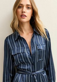 Blonde vrouw in een marineblauwe, lange mouwen, knoopsluiting jurk met witte verticale strepen en een gestrikte tailleband, staand tegen een neutrale achtergrond.