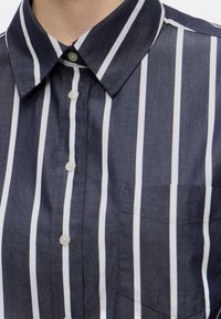 Gros plan d'une personne portant une chemise bleu foncé à rayures blanches verticales et boutons blancs, avec un col pointu et une poche poitrine.