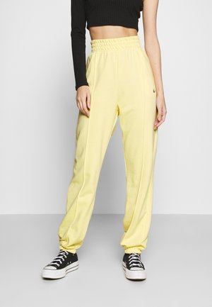 Personne portant un pantalon de jogging jaune taille haute, un top noir court à manches longues et des baskets noir et blanc, debout devant un fond uni.