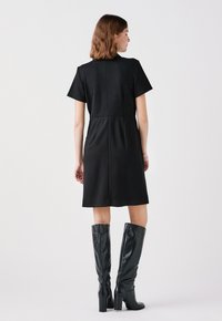 Schwarzes kurzärmeliges Kleid mit taillierter Silhouette, glattem Stoff und knielangem Design, kombiniert mit glänzenden schwarzen knielangen Stiefeln mit Absatz.