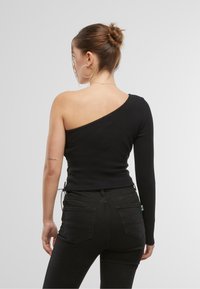 Svart én-skulder topp med lang arm på den ene siden, med en tilpasset design og rynkedetaljer langs siden, i kombinasjon med svarte jeans.
