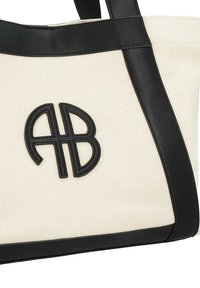 Sac fourre-tout en toile crème avec des accents et des poignées en cuir noir. Présente un logo noir brodé "AB" sur le devant. Forme rectangulaire.