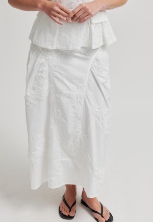 Kobieta ubrana w białą, haftowaną spódnicę i pasujący top, z splecionymi rękami i czarnymi klapkami na stopach, stojąca na białej podłodze.