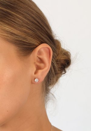 Runde silberne Steckerohrringe mit klaren Steinen, die eine glatte metallische Oberfläche und eine sichere Befestigung aufweisen, getragen im Ohrläppchen.