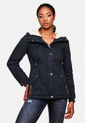 Femme portant une veste d'hiver noire à capuche avec doublure en fourrure et poches avant, associée à un jean bleu foncé délavé.