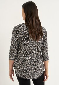 Zwarte blouse met een crèmekleurig bloemenpatroon, driekwartmouwen, ronde zoom en lichte stof. Draag met zwarte broek.