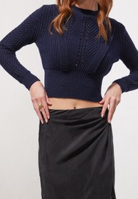 Pull court en maille bleu marine avec texture côtelée, présentant des lignes verticales et des mailles ajourées. Associé à une jupe noire drapée.