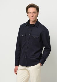 Camicia button-up blu scuro realizzata in tessuto morbido, con colletto classico, due tasche sul petto e dettagli in metallo sui bottoni.
