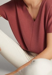 Karmelowy sweter z dekoltem w kształcie litery V, krótkimi rękawami i przeszyciami, zestawiony z obcisłymi kremowymi spodniami. Gładka tekstura.