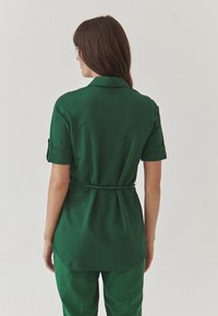 Cămașă verde dintr-un material ușor, cu guler, mâneci scurte rulată și talie cu curea, având spatele ușor prelungit.