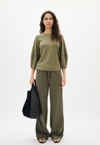Olivgrünes, lässig geschnittenes Oberteil mit 3/4-Ärmeln, kombiniert mit weiten olivfarbenen Hosen. Eine schwarze Tragetasche und Flip-Flop-Sandalen runden das Outfit ab.