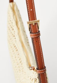 Beige woven bag with brown faux-leather strap, gold buckle, and stitching details. Features a zipper closure for secure access.