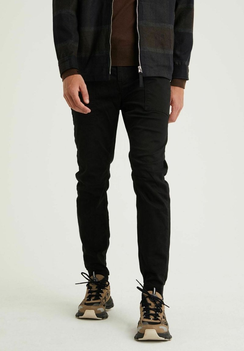 Homme portant un pantalon noir slim, des baskets marron et noir, et une veste zippée foncée avec des manches marron, debout sur un fond uni.