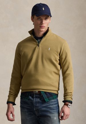 Junger Mann trägt einen hellbraunen Pullover mit Viertel-Reißverschluss, ein blau-grün kariertes Hemd, blaue Jeans, eine marineblaue Mütze und eine Armbanduhr, steht vor einem schlichten Hintergrund.