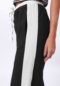 Schwarze Sporthose mit einem weißen Seitenstreifen, elastischem Bund mit Kordelzug und Seitentaschen. Glatte Stofftextur mit einer lockeren Passform.