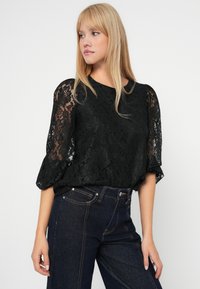 Czarna koronkowa bluzka z rękawami o długości trzech ćwierci z bufiastym wykończeniem, ozdobiona teksturowanym kwiatowym wzorem. Połączona z ciemnoniebieskimi dżinsami o wysokim stanie.