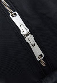 Tissu en nylon noir avec une fermeture éclair métallique argentée dotée d'un tirette rectangulaire et de l'étiquette "Carhartt" gravée dessus.