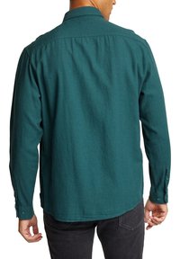 Camisa de manga larga color azul verdoso con cuello abotonado, textura suave y pliegues en la espalda, llevada sobre pantalones oscuros. Puños con botones en las muñecas.