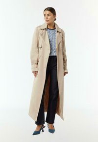 Beige Trenchcoat mit tailliertem Gürtel, doppelter Knopfleiste und breiten Revers. Tragen über einem gemusterten Oberteil, kombiniert mit dunklen Jeans und High Heels.