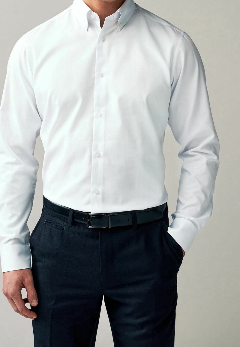 Chemise blanche à col, associée à un pantalon formel sombre et une ceinture noire. Le tissu semble lisse avec une coupe ajustée.