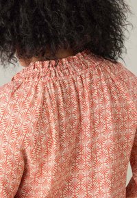 Blusa a fantasia floreale in corallo e crema, con scollo arricciato e tessuto testurizzato, riportante intricate decorazioni a forma di foglia.