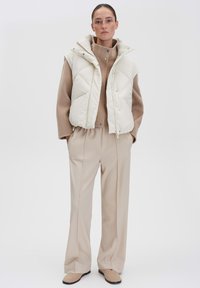 Weiße gepolsterte Weste mit hohem Kragen, getragen über einem beigen Pullover und beigen weiten Hosen, was eine stimmige neutrale Farbpalette schafft.