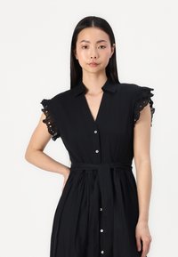 Melnas kleitas ar īsām frillētu piedurknēm, pogām priekšā un saitēta josta. Izgatavota no viegla auduma ar gludu tekstūru.