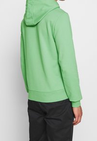 Felpa con cappuccio verde in tessuto morbido, con tasca a marsupio e polsini a costine. Mostrata dal retro, abbinata a pantaloni neri.