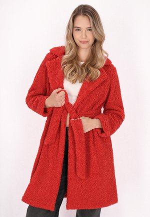 Cappotto rosso, texturizzato e peloso, con cappuccio, apertura frontale e cravatta abbinata. Presenta maniche lunghe e arriva fino a metà coscia. Abbinato a una maglietta bianca.