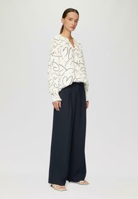 Blouse blanche à motif de cœurs noirs, manches longues avec des poignets froncés, associée à un pantalon navy à jambes larges et des sandales blanches à brides.
