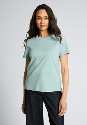 Frau mit langem dunklem Haar, die ein hellblaues Kurzarmhemd und dunkelblaue Jeans trägt, steht vor einem schlichten hellen Hintergrund.